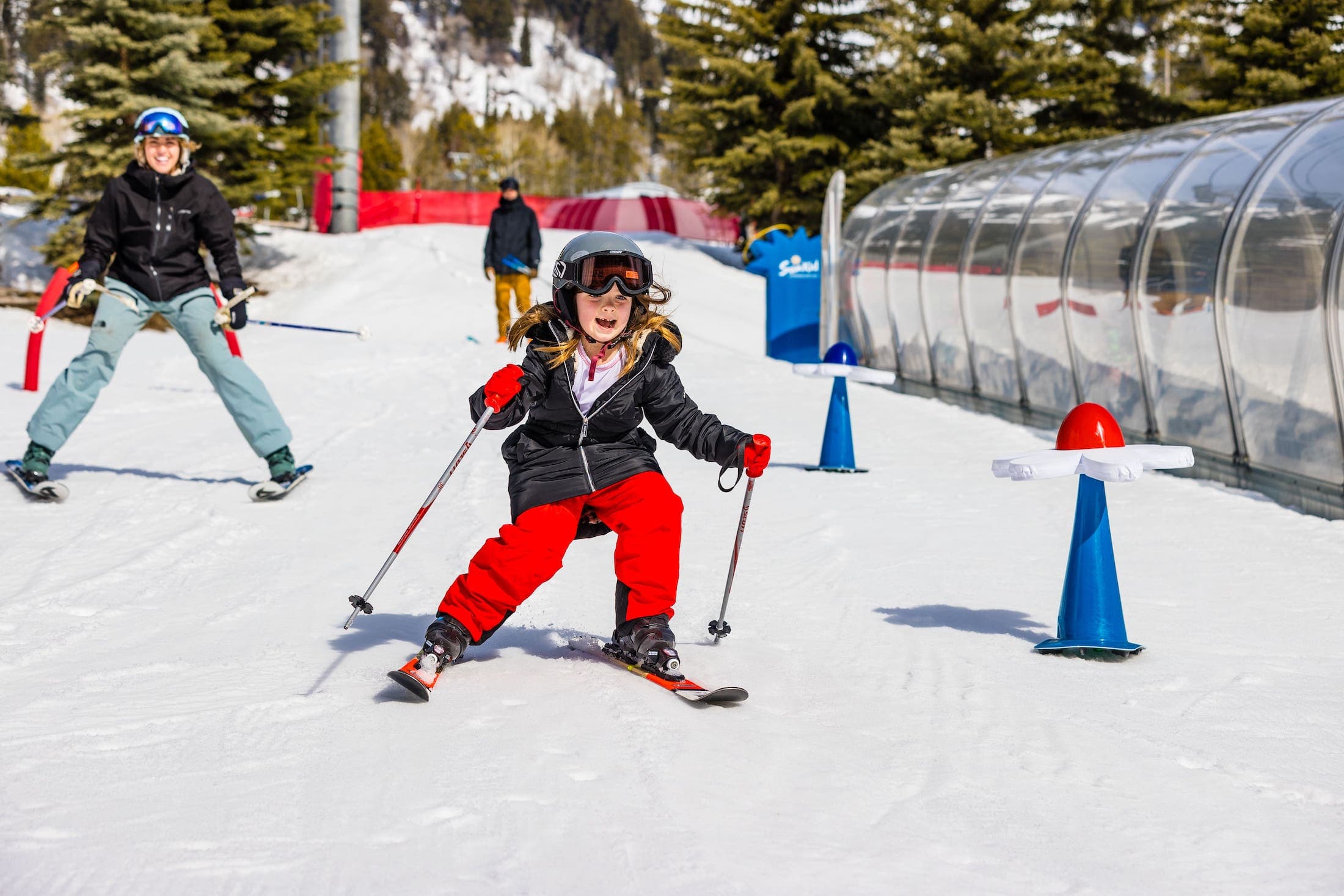 A photo of a child skiing down a slope being taught and monitored by a man. Another person can be seen in the background.