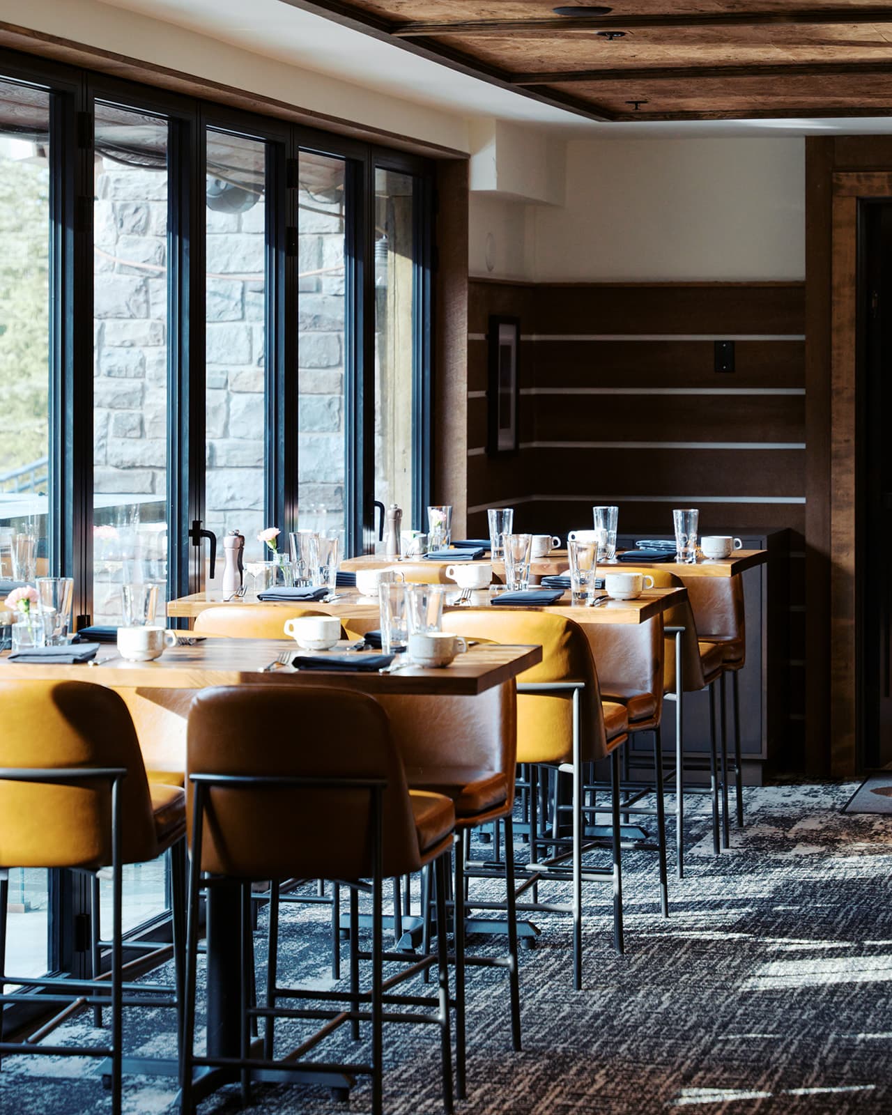 Multiple restaurant tables with seating and place settings next to a window, with the sun streaming in from the left