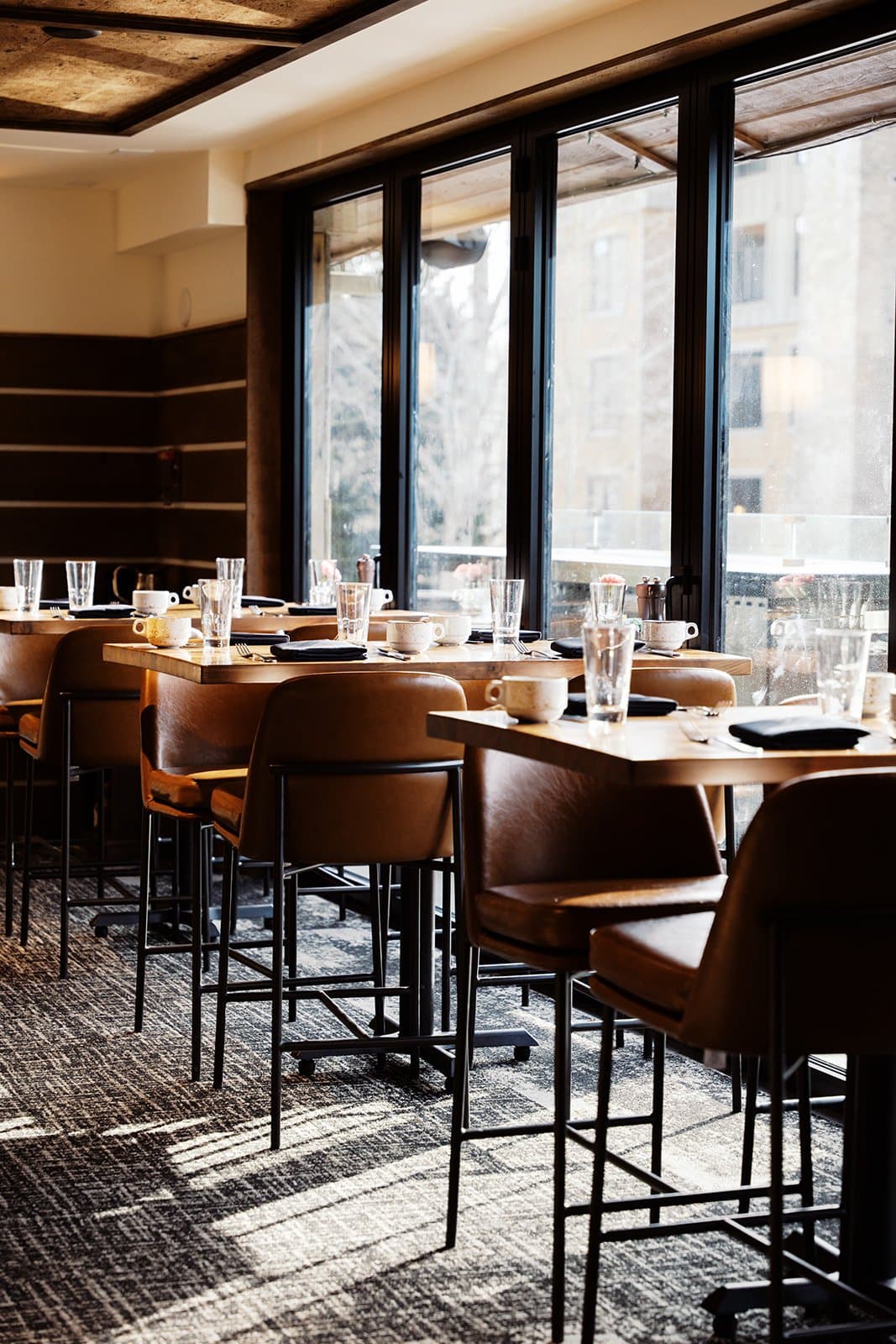 Three restaurant tables next to a lit window with four seats each, prepared with place settings