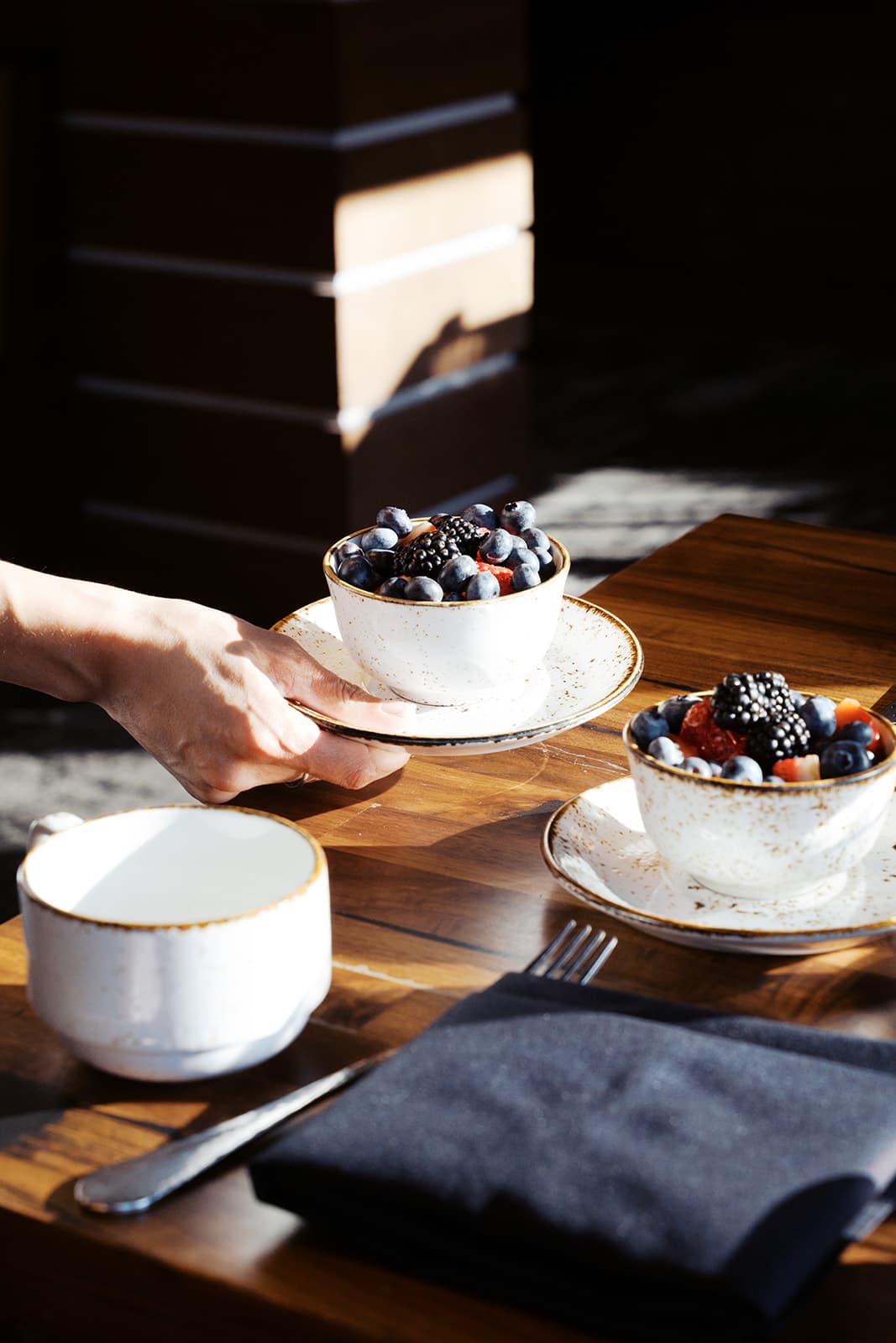 A person setting down a bowl of mixed blueberries, strawberries, and blackberries onto a table next to another bowl of mixed berries.