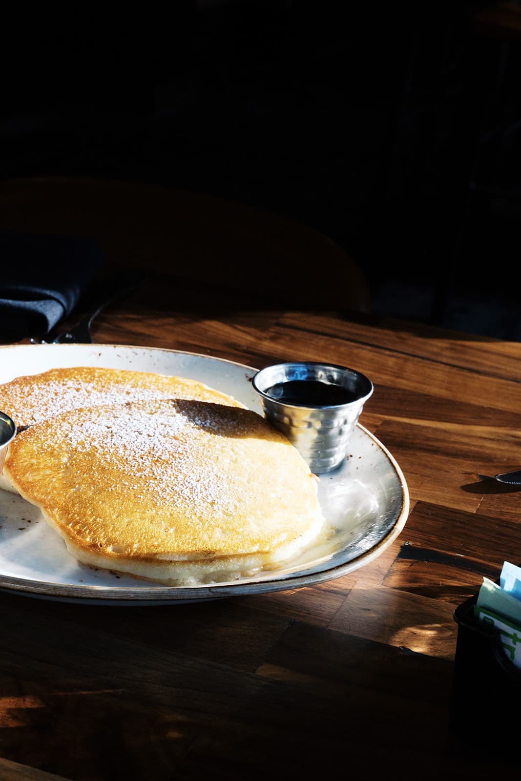 A plate of fluffy pancakes dusted with powdered sugar and maple syrup in a silver dip dish.