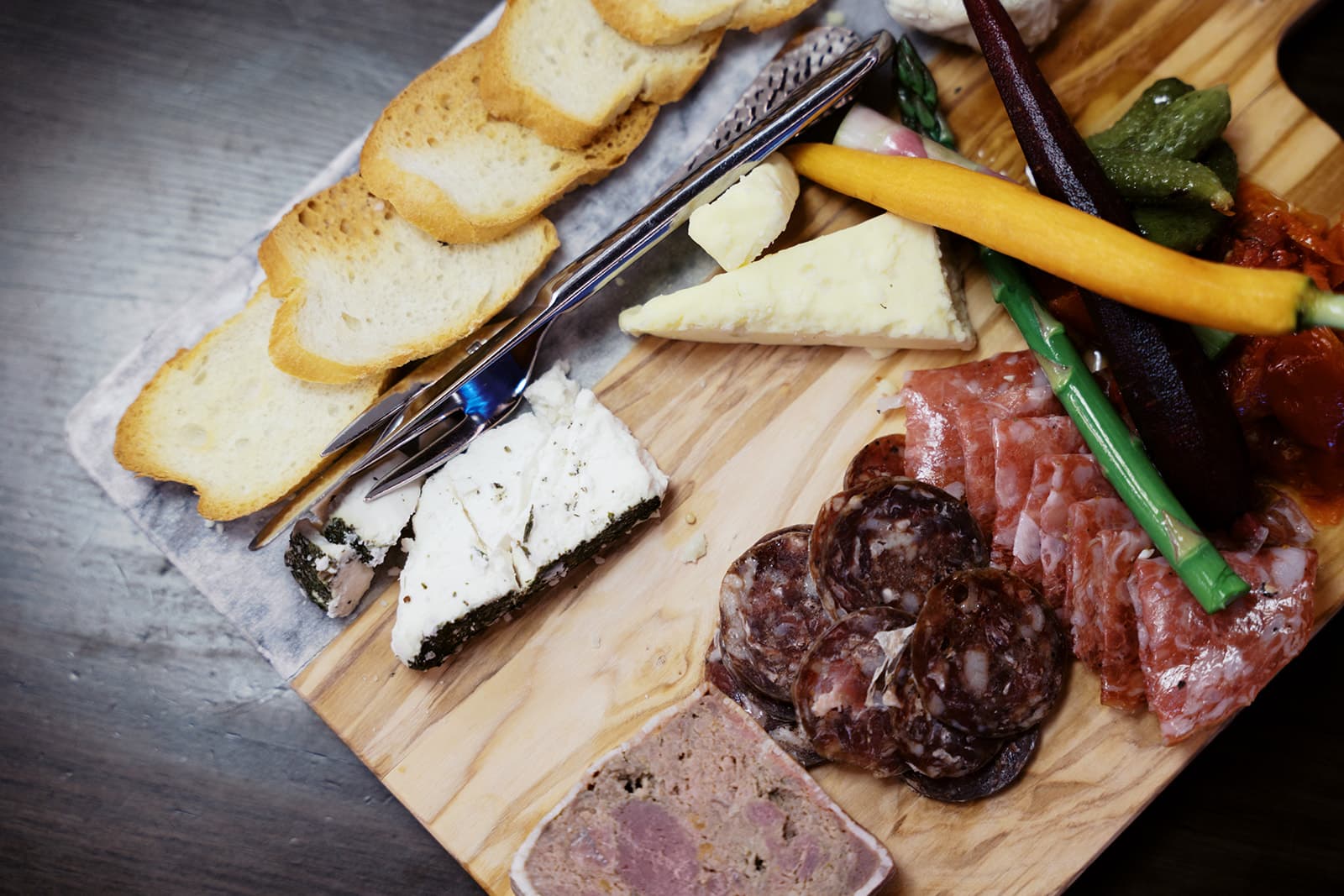 Closeup image of a mixed charcuterie board containing three varieties of charcuterie meat, two cheeses, crispy baguette crackers and variety of pickled veggies