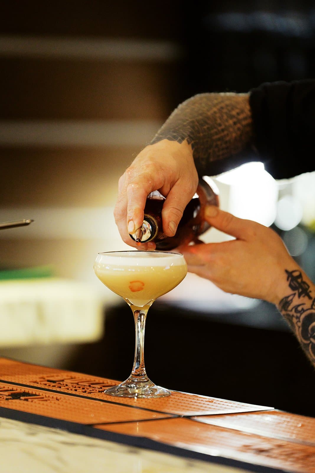 A bartender pouring a yellow orangish toned drink into a le coupe glass.