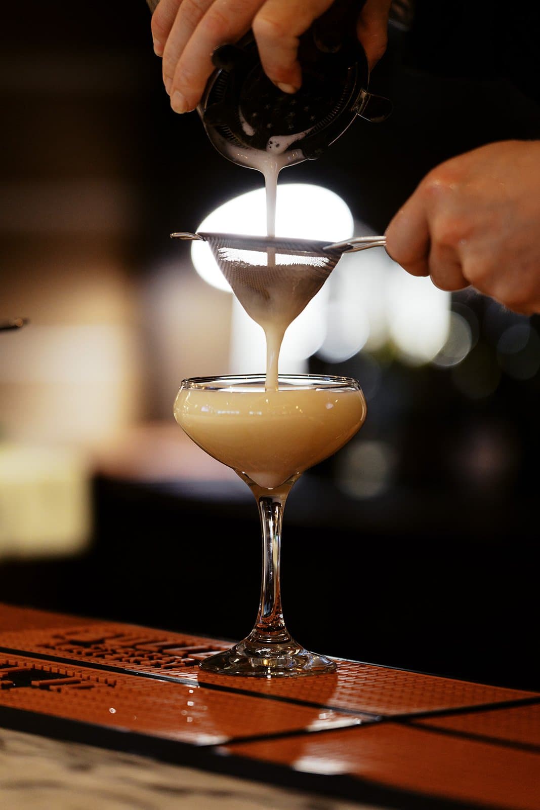 A bartender pouring a yellow orangish toned drink into a le coupe glass through a sifter.