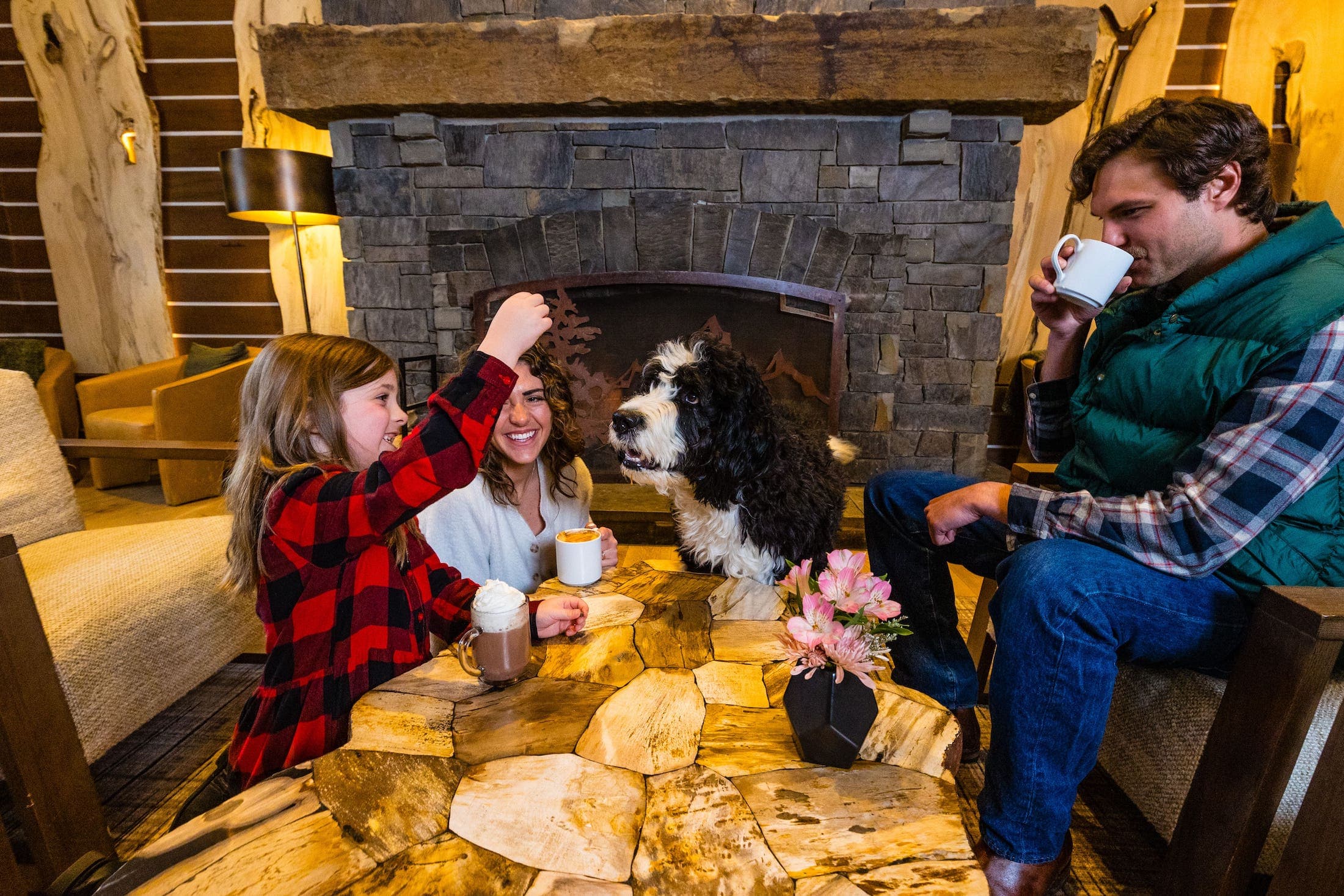 A family enjoys hot drinks at front of a fireplace as a young girl feeds their dog a treat