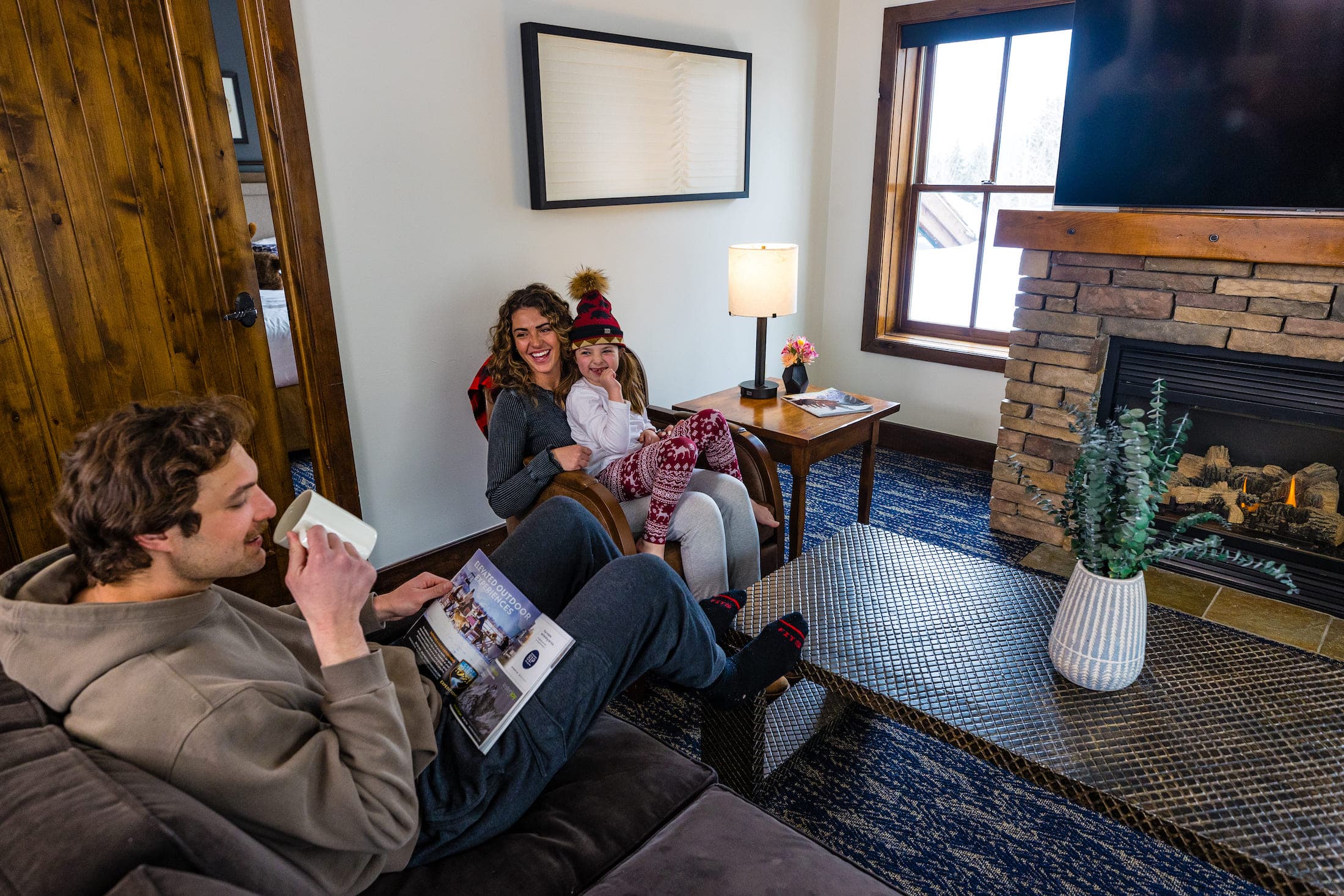 A family consisting of a man, woman, and child sit in a lounge next to a door. There is a fireplace opposite to them. The woman is reading a magazine or brochure while the woman holds the child on her lap, who is wearing a winter hat.