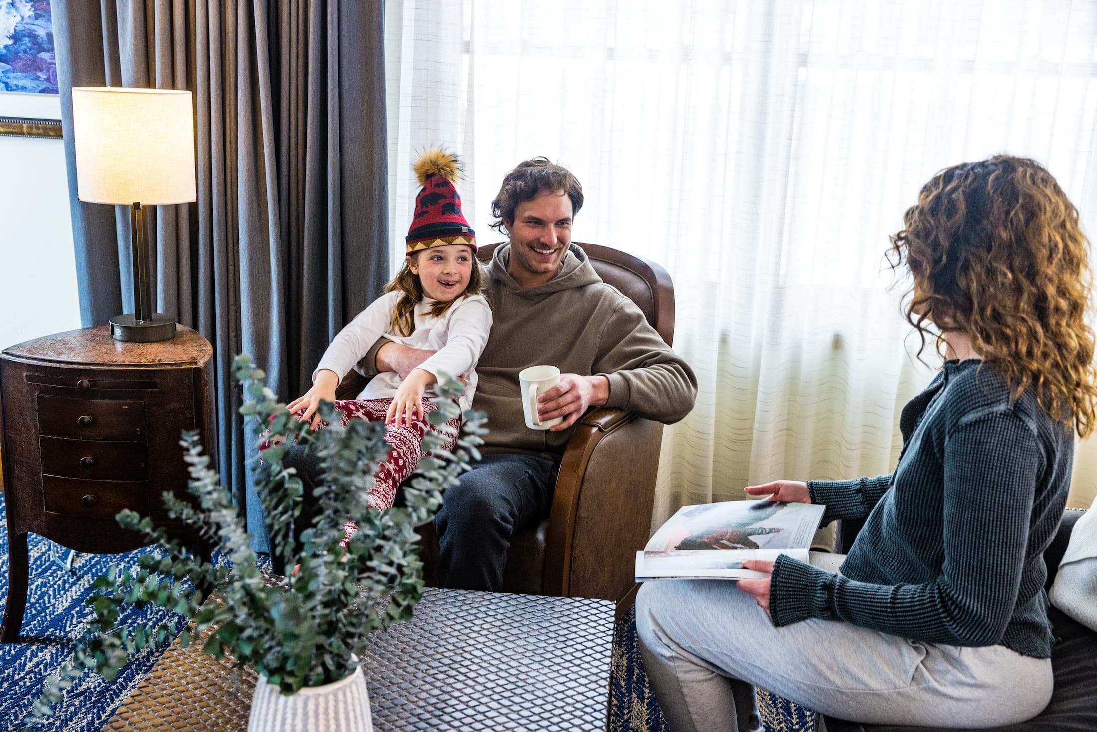 A family consisting of a man, woman, and child sit in a lounge backlit by a window. The woman is reading a magazine or brochure while the man holds the child on his lap, who is wearing a winter hat.