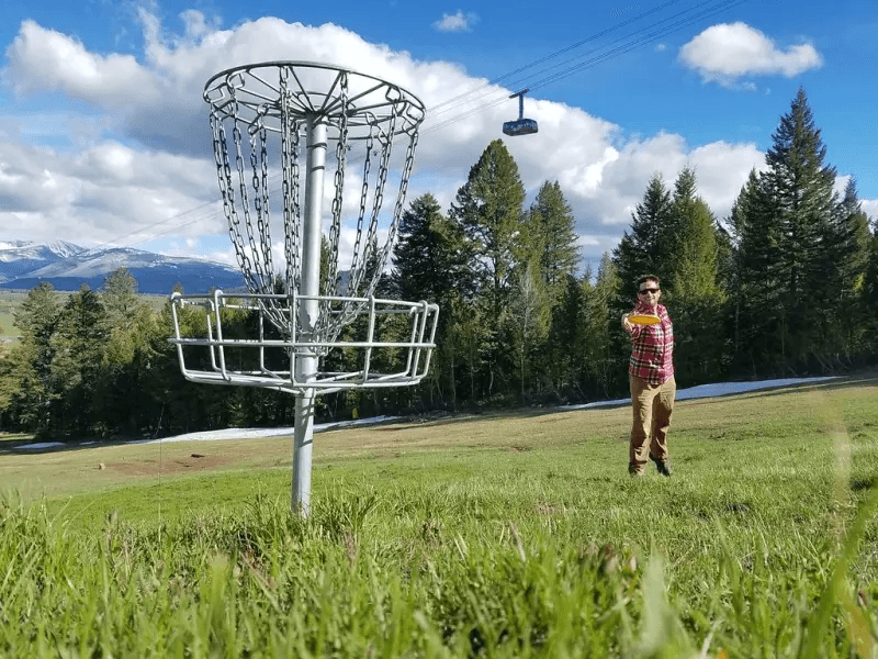 A man plays Disc Golf in a field with mountains peaking behind trees, high above him is a gondola lift