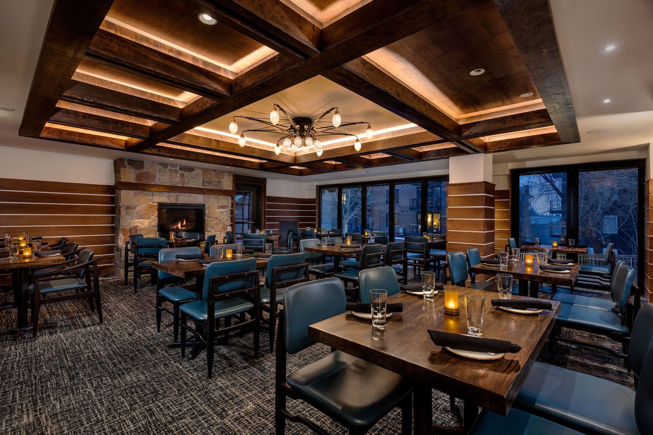 The Spur Restaurant dining room, in the evening. Multiple tables with place settings can be seen as well as a fireplace on the back wall and windows to the side
