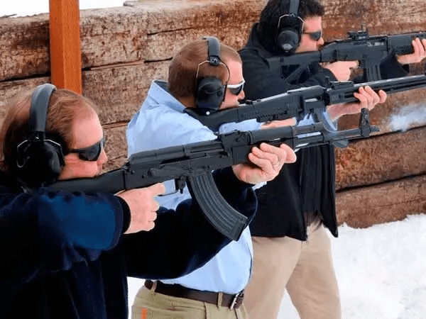 Three men at a shooting range with ear and eye protection holding rifles, one rifle has smoke coming from its barrel