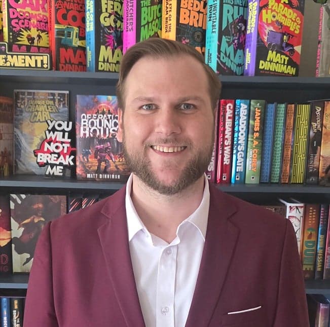 Jordan Mayo in a maroon blazer and white shirt smiling in front of a bookshelf of science fiction novels