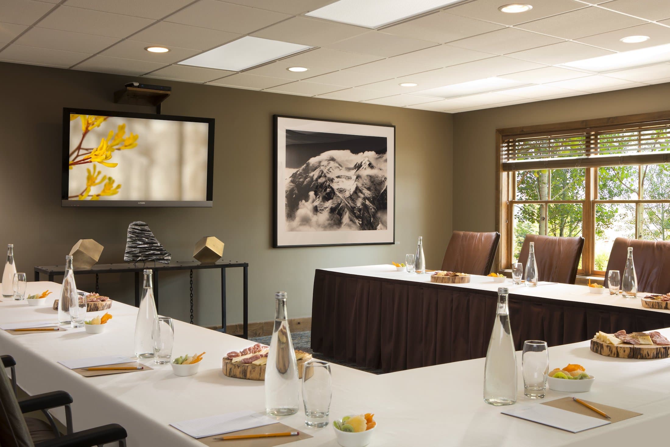 A photo of the Teton Moutain Lodge Four Pines Meeting Room. A U-shaped table can be seen with water glasses, bottles of water, and dishes of fresh fruit. Two large paintings can be seen hanging on the walls.