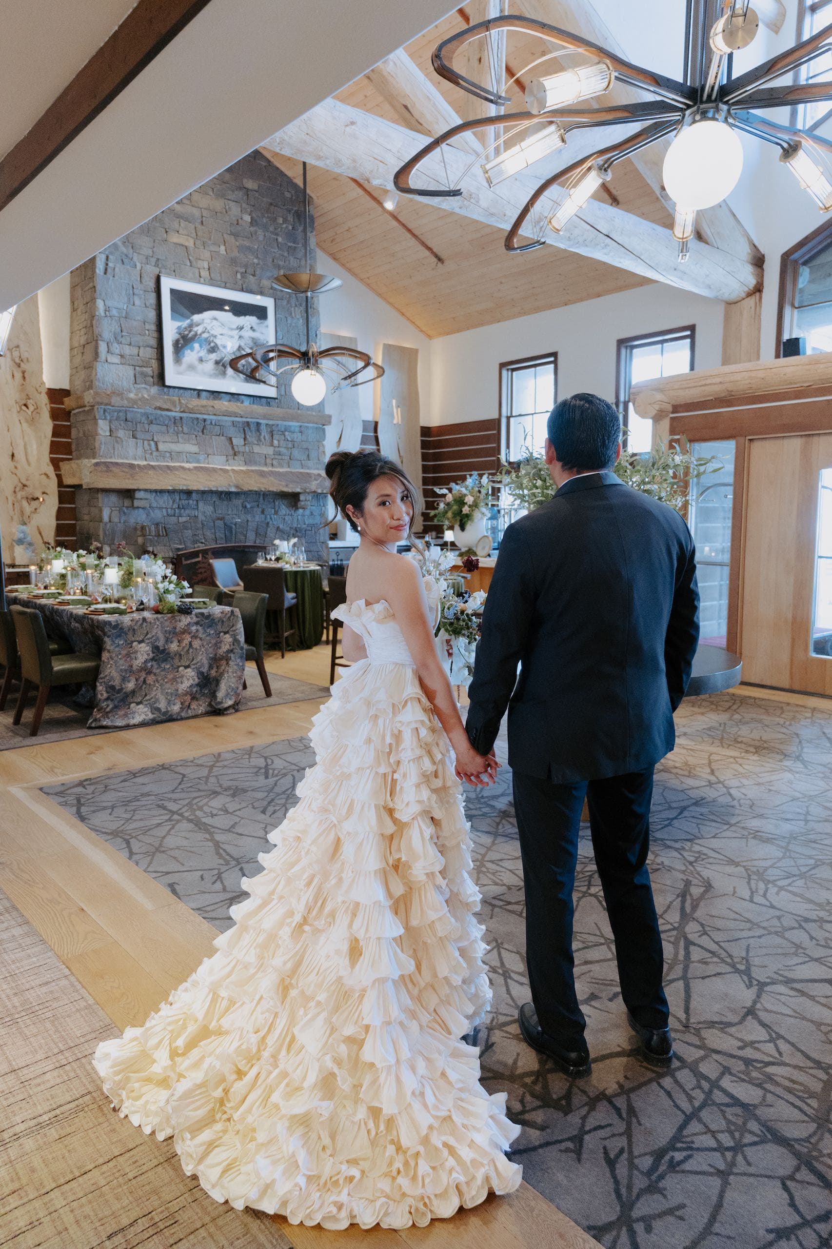 A wedded couple standing in a reception room, the man is facing away while the woman looks behind her at the camera