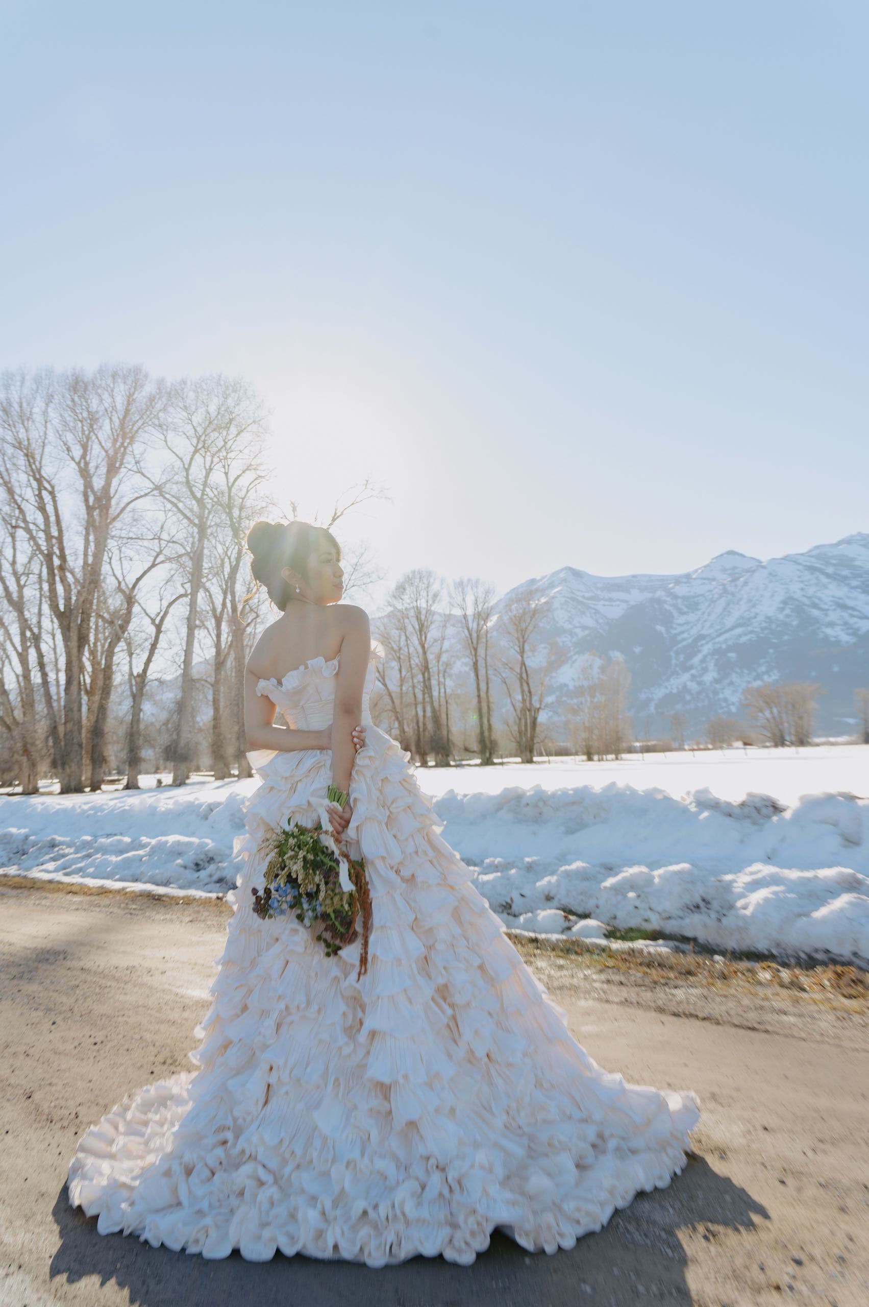 A photo of a bride in a wedding dress standing outside backlit by the sun