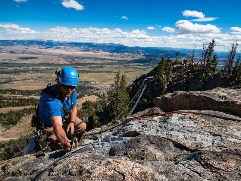 A man wearing climbing gear climbs up a granite cliff amidst the backdrop of a blue sky and vast landscape. A rope bridge can be seen spanning from a wooded steep hill