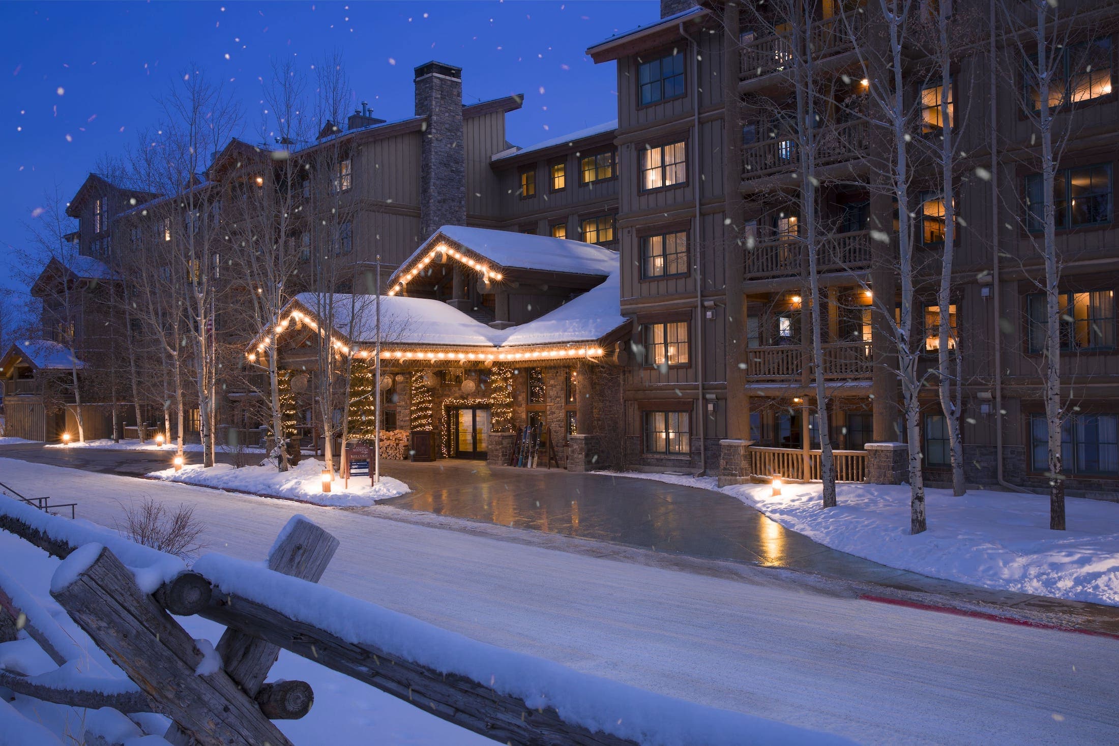 The Teton Mountain Lodge entrance in the evening across from a wooden fence, while snow lightly falls